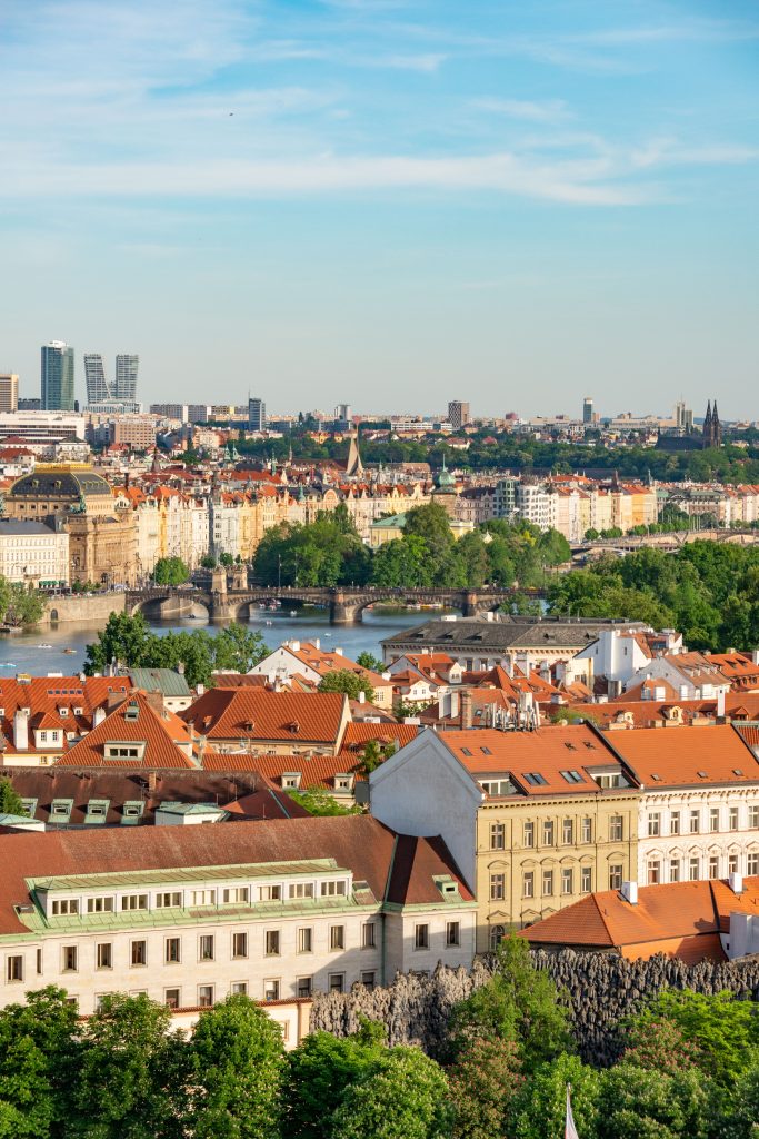 A stunning aerial view of historic Prague with red rooftops and lush greenery under a clear blue sky.